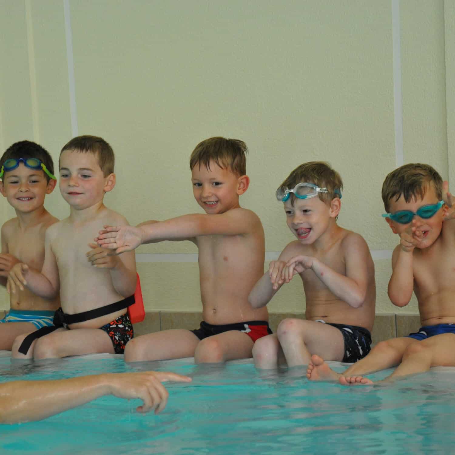 Auf dem Bild sind fünf Kinder bei einem Anfänger-Schwimmkurs zusehen. Sie sitzen am Beckenrand und machen die Übung nach, die ihnen vom Schwimmlehrer vorgezeigt wird.